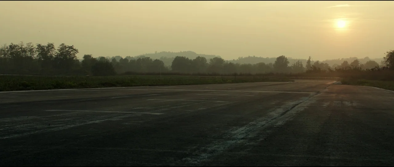 Ferrari (2023) movie still: establishing — A wide view of an empty asphalt runway extending into a hazy rural landscape. Dense green trees line the…