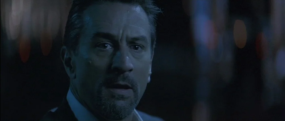 Heat (1995) movie still: closeup — A night time city street at street level with dark, blurred background lights. A man with a dark suit and goatee…
