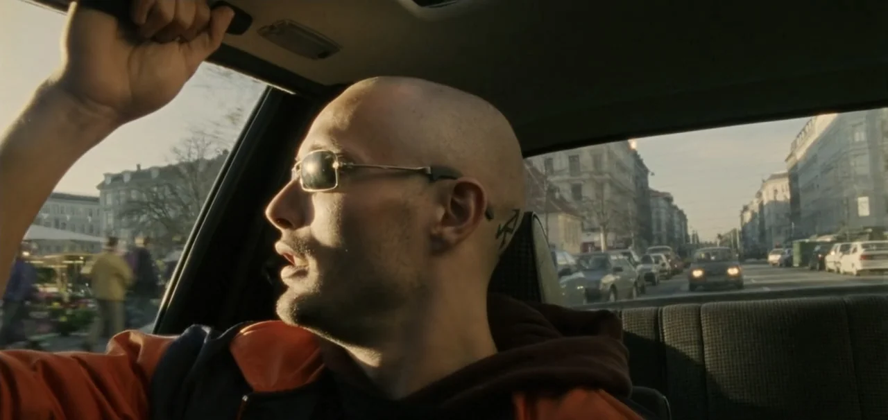 Pusher (1996) movie still: medium — A man rides in the passenger seat of a car traveling through a city. Outside the window, urban street scenes…