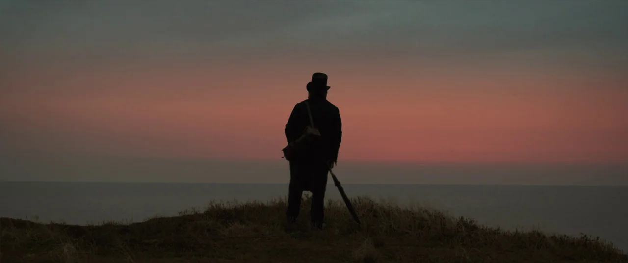 Mr. Turner (2014) movie still: fullbody — A grassy cliff top overlooks a calm ocean at sunset. A man wearing a top hat stands at the edge while…