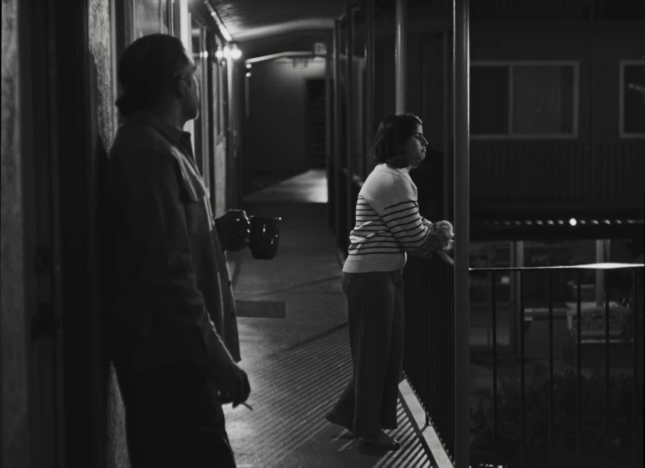 Fremont (2023) movie still: medium — An outdoor motel walkway at night with metal railings and concrete floors. A man and a woman stand apart on the…
