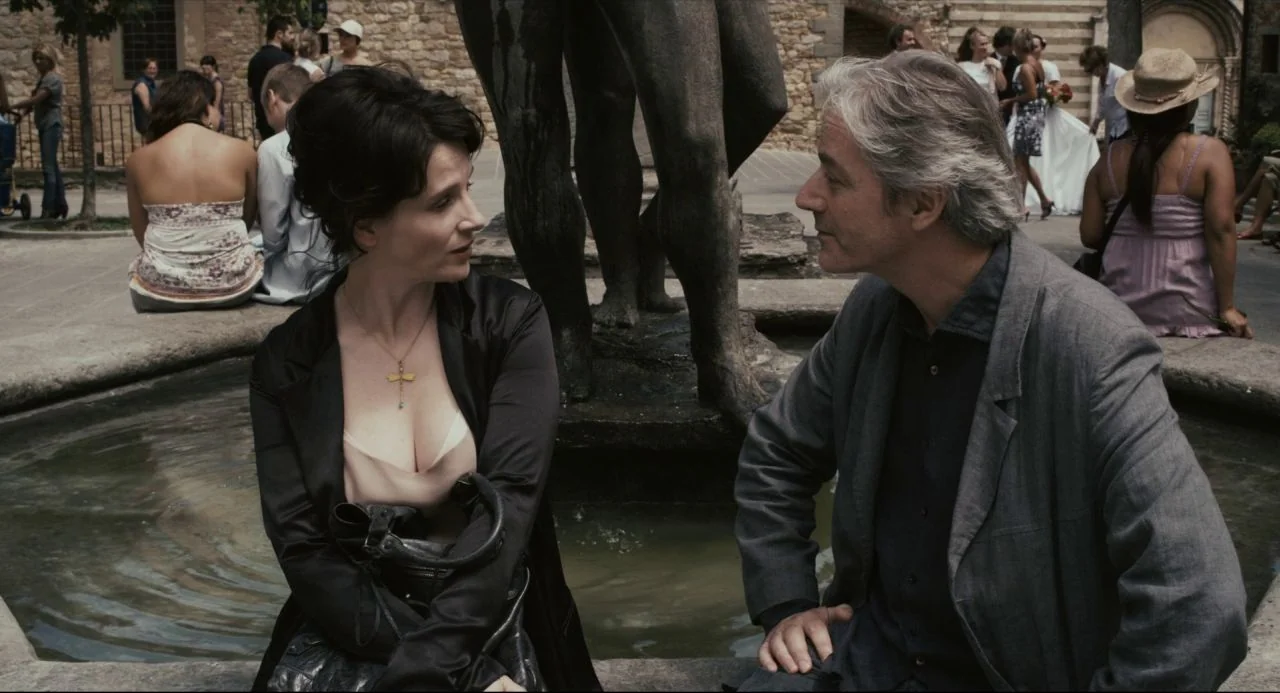 Certified Copy (2010) movie still: medium — A man and woman sit opposite each other on the stone rim of a fountain. Other people are visible in the…