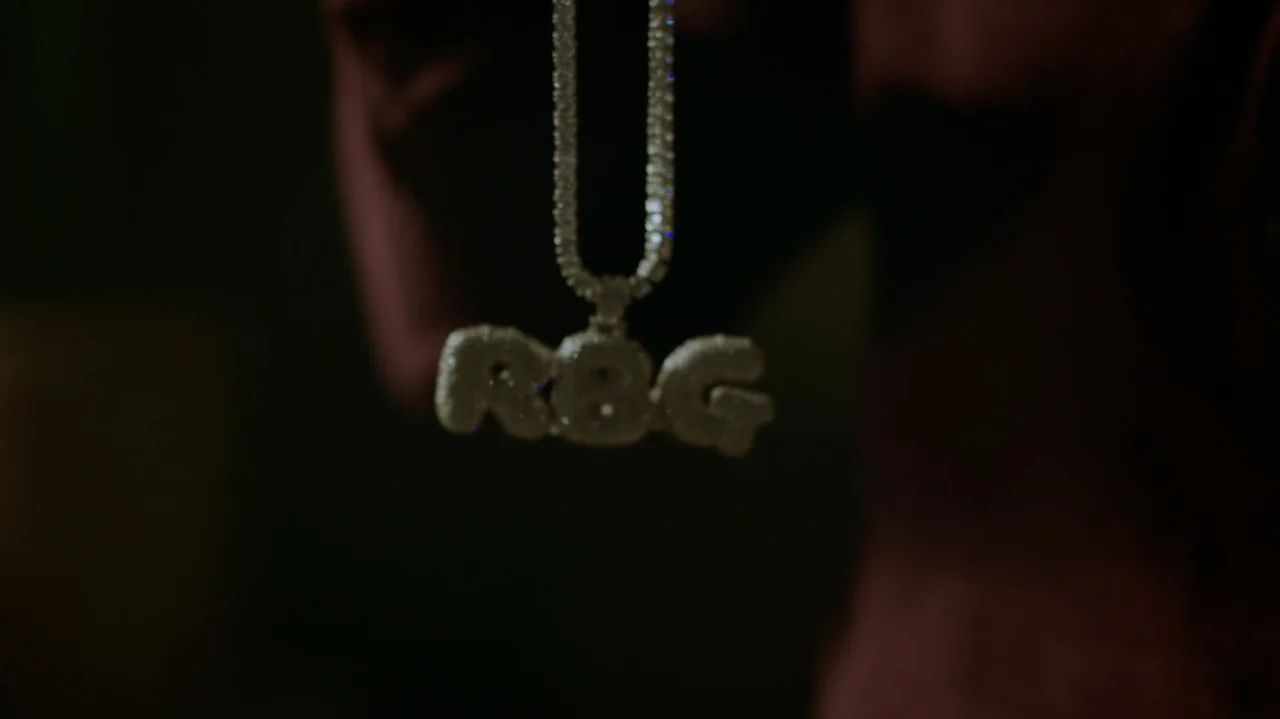 Die Hart (2023) movie still: closeup — A close-up view of a sparkling, diamond-encrusted pendant hanging from a thin chain. The letters R, B, and G…