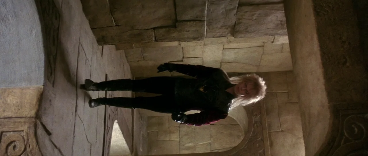 Labyrinth (1986) movie still: fullbody — An interior stone corridor features block-like masonry walls and arched doorways. A man with voluminous…