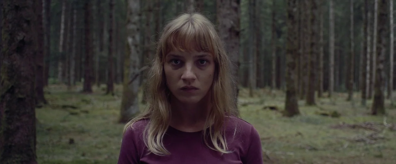 Meanwhile On Earth (2024) movie still: medium — A woman stands in a densely packed pine tree stand. She stares directly into the lens while…
