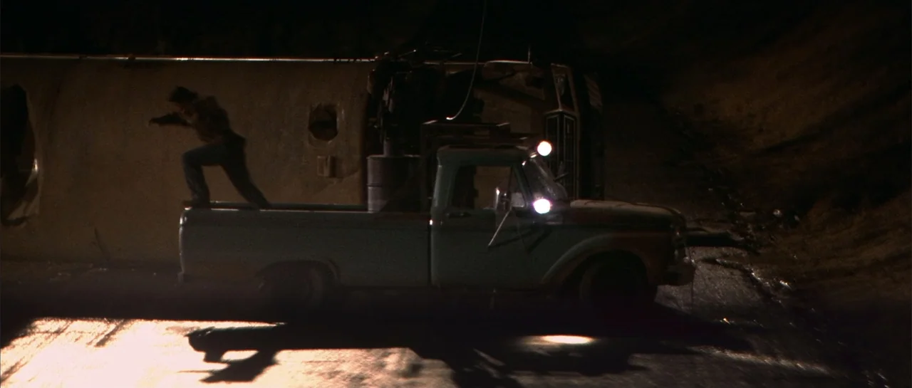 Jeepers Creepers 2 (2003) movie still: wide — A dark rural road at night features a light blue pickup truck parked prominently. A man stands in the…