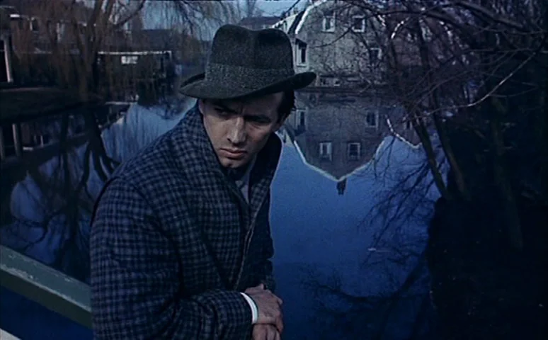 Mill of the Stone Women (1960) movie still: medium — A man in a fedora stands on a bridge over a dark canal. The reflection of a building appears on…