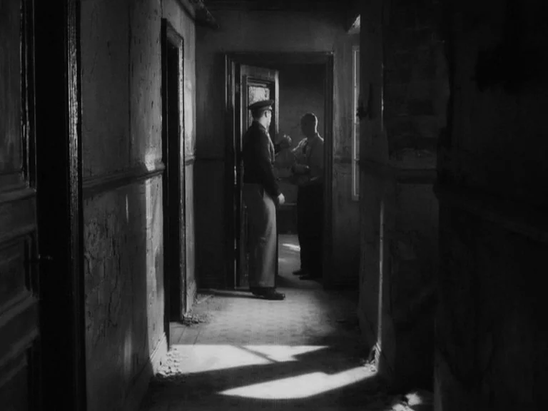 The Good German movie still: wide — A dark, decaying interior hallway shows worn paint and shadows on the walls. Two men stand in an open doorway at…