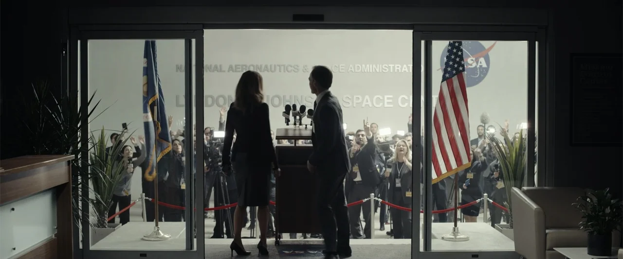 Moonfall (2022) movie still: fullbody — A modern lobby with glass doors overlooks a large press room with signage. A man and woman in business…