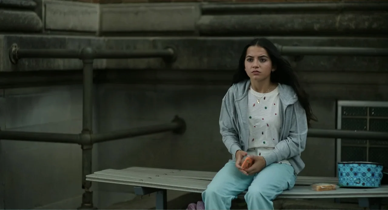Turtles All the Way Down (2024) movie still: medium — A woman sits on a metal bench outdoors against a dark stone wall. She holds a small orange…