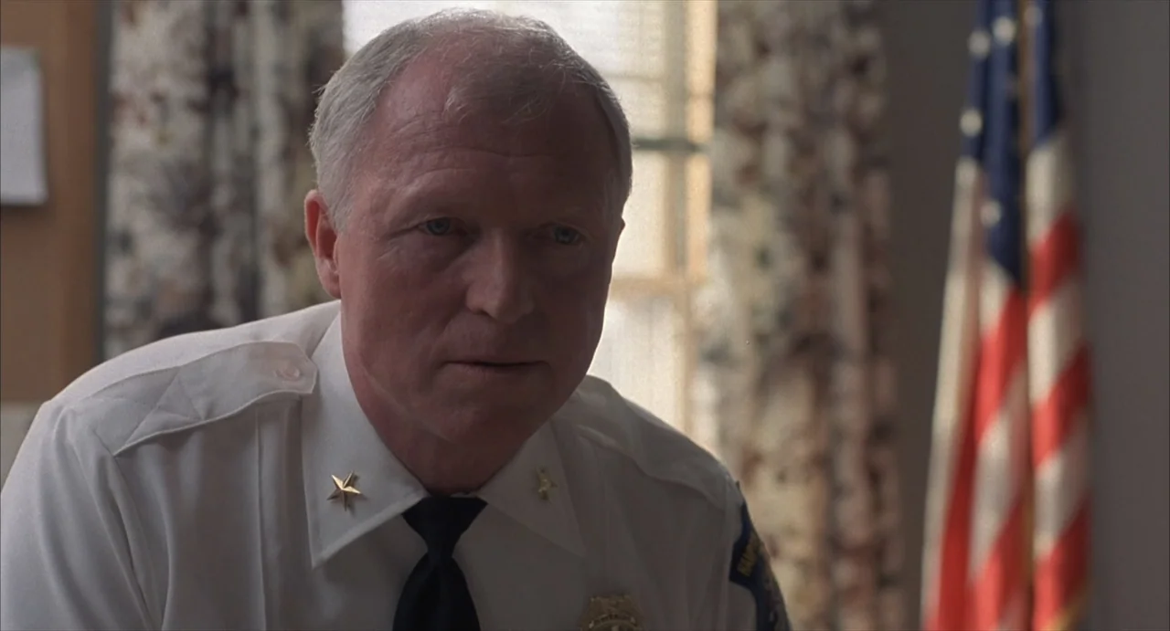 Masquerade (2023) movie still: medium — An office interior features a wooden paneled wall and a blurry American flag backdrop. A middle-aged man in…