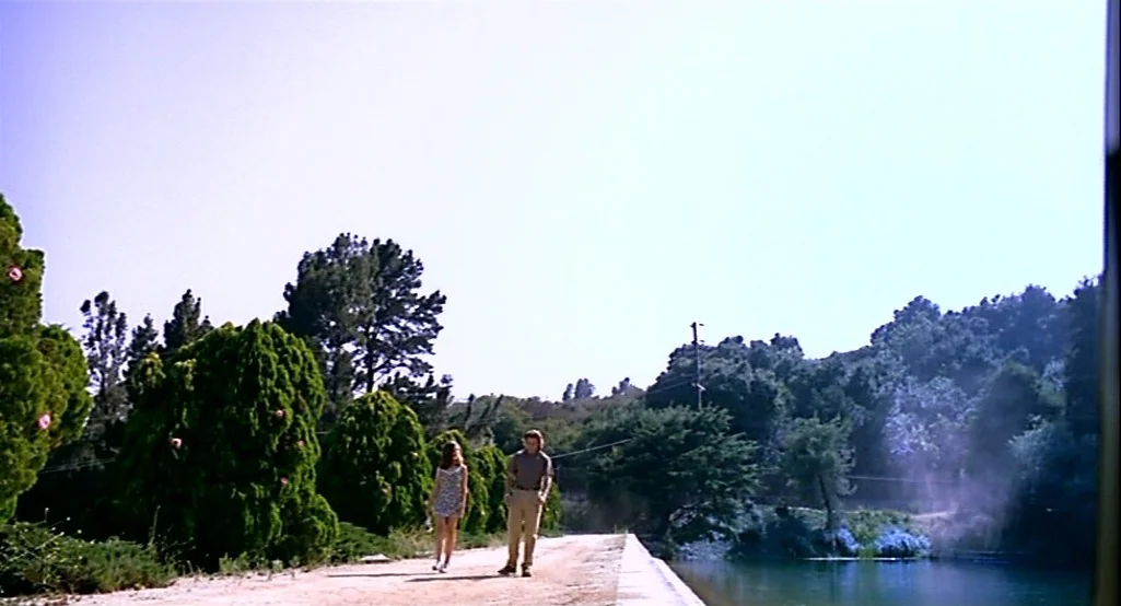 Nowhere (1997) movie still: wide — A dirt path runs alongside a river under a clear, bright sky. A man and woman walk toward the camera while…
