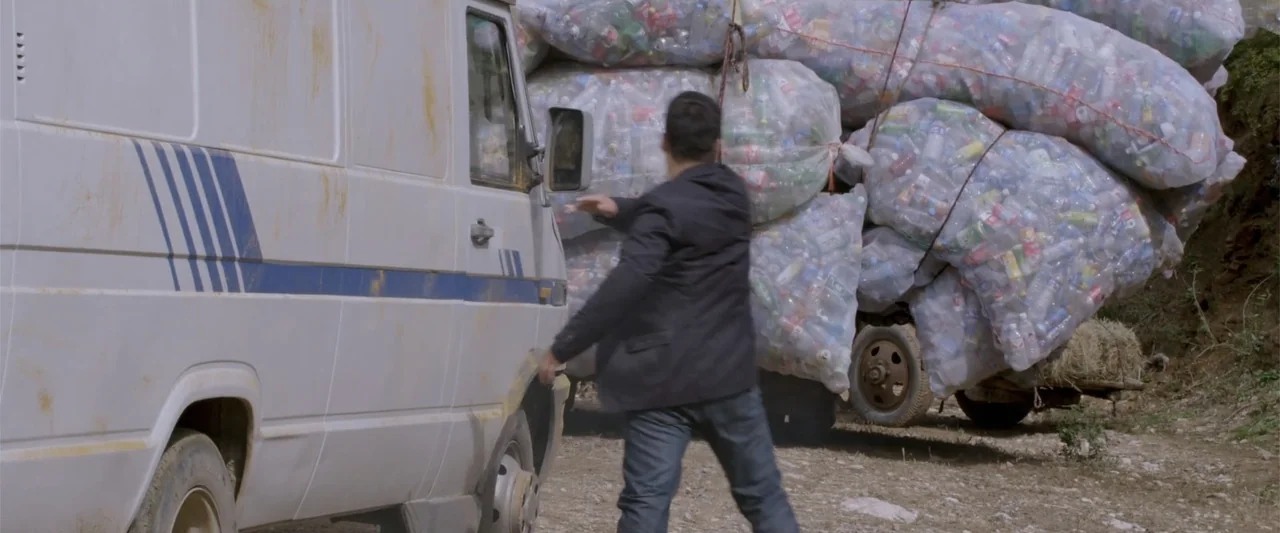 Skiptrace (2016) movie still: medium — A dirty white cargo van is parked on a dusty dirt road. A man in a dark jacket reaches toward the van door…