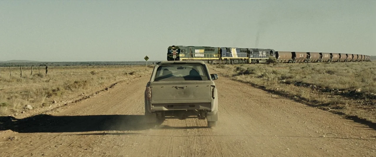 The Rover (2014) movie still: wide — A flat, arid landscape stretches toward the horizon under a clear, pale sky. A dusty pickup truck approaches a…