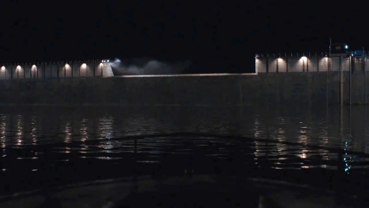 Night Moves (2013) movie still: establishing — A massive concrete dam structure spans across a dark expanse of water at night. Rows of lights are…