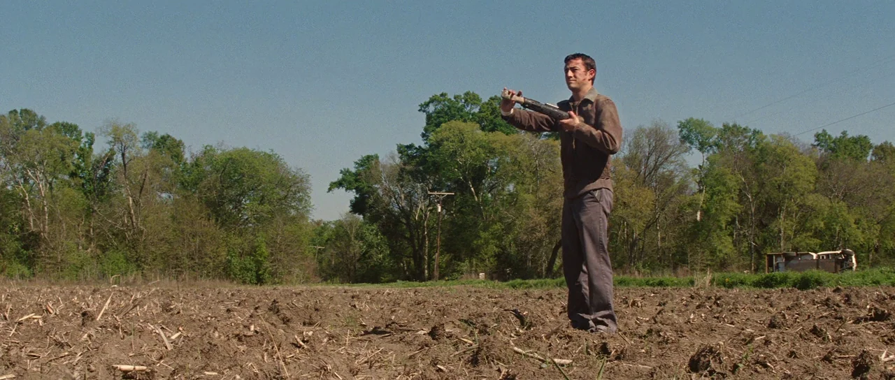 Looper (2012) movie still: fullbody — A man stands in a vast, dry agricultural field during the middle of the day. He holds a firearm while looking…