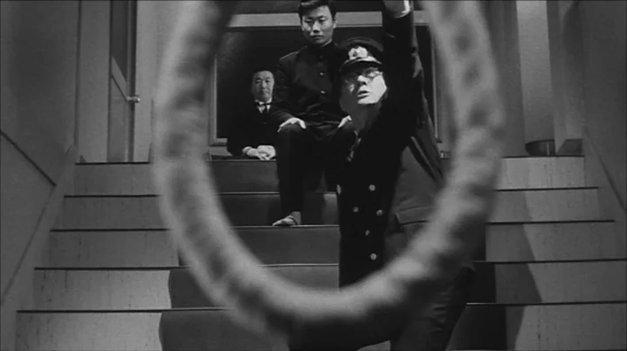 Death By Hanging (1968) movie still: medium — Three men stand on a staircase with a blurry rope in the immediate foreground. The foreground rope…