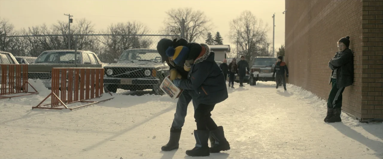 The Best Christmas Pageant Ever (2024) movie still: fullbody — A snow-covered school parking lot features a chain-link fence and parked vintage…