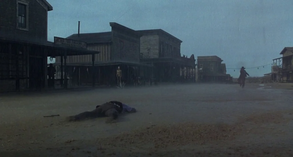 The Quick and the Dead (1995) movie still: wide — A rain-swept western main street is lined with weathered wood buildings under gray skies. A man…