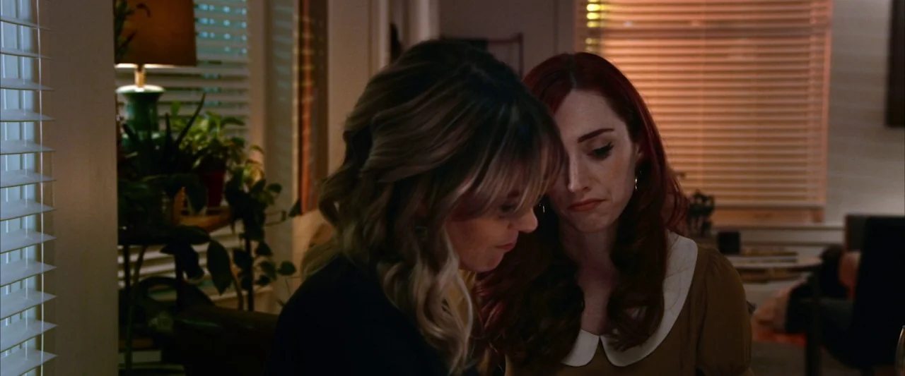 The Stylist (2020) movie still: medium — A furnished interior space with window blinds and a small houseplant on a stand. Two women stand close…