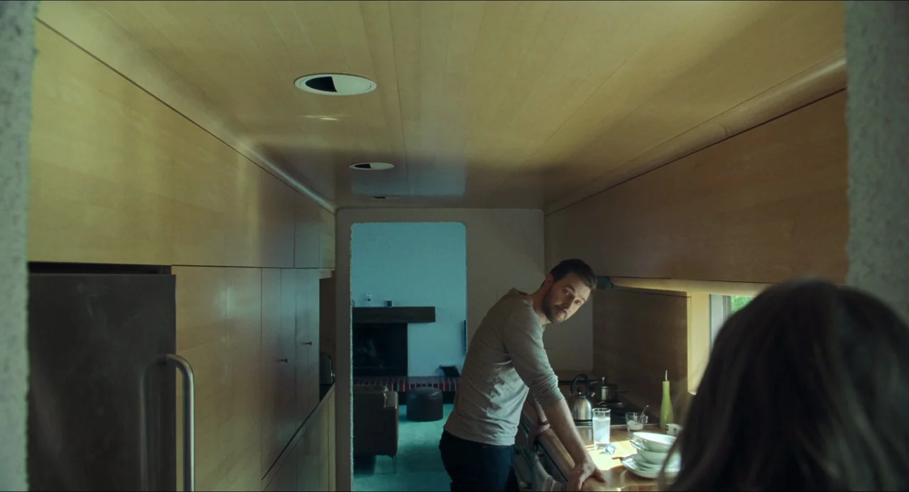 The Lodge (2019) movie still: medium — A modern kitchen with extensive wood cabinetry and a view of an adjacent room. A man stands by the counter…