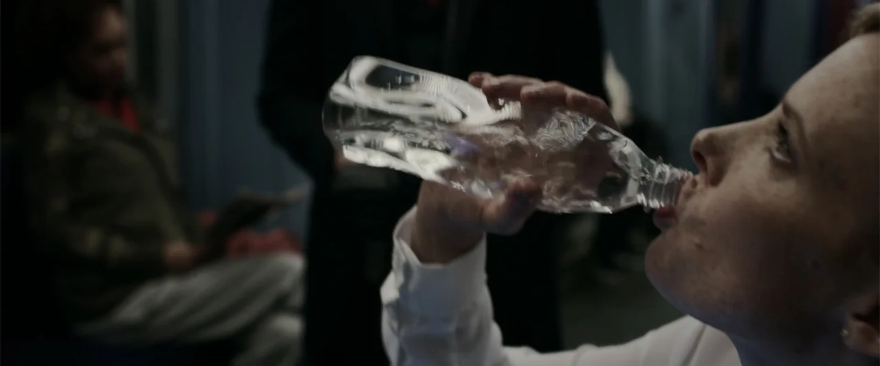 Howl (2015) movie still: closeup — A dimly lit subway car contains a passenger drinking from a plastic bottle. A woman in a white shirt drinks water…