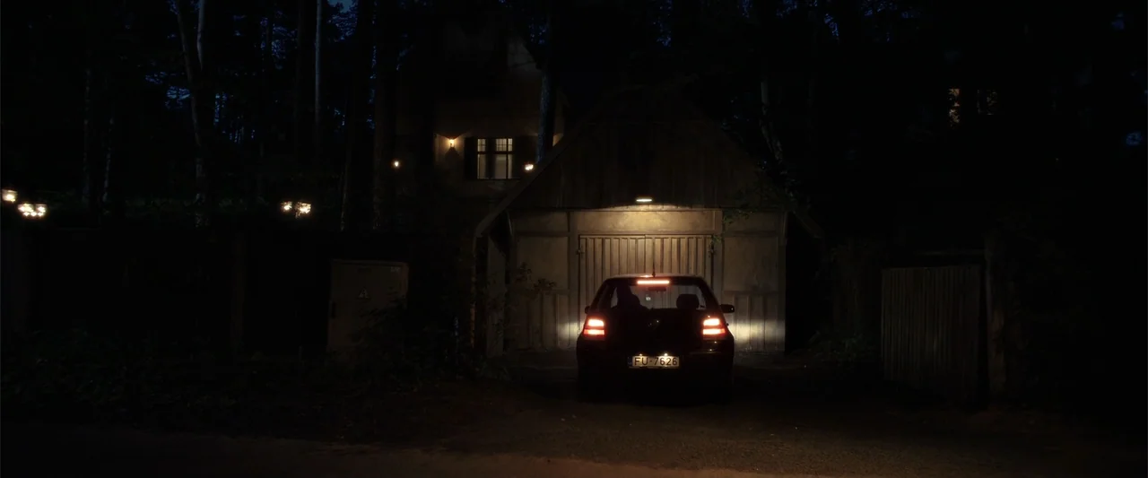 The Good Neighbor (2022) movie still: wide — A dark car is parked in a tree-lined driveway in front of a wood-paneled garage. The vehicle has its…