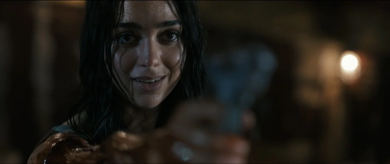 Scream VI (2023) movie still: closeup — A dark, dimly lit interior space with an indistinct background. A young woman with damp hair and a stained…
