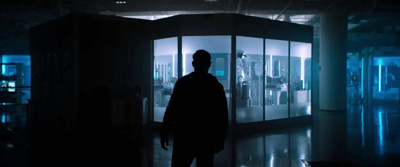 Venom (2018) movie still: fullbody — A dark industrial laboratory contains a series of glass-walled rooms illuminated by blue lights. A lone man…