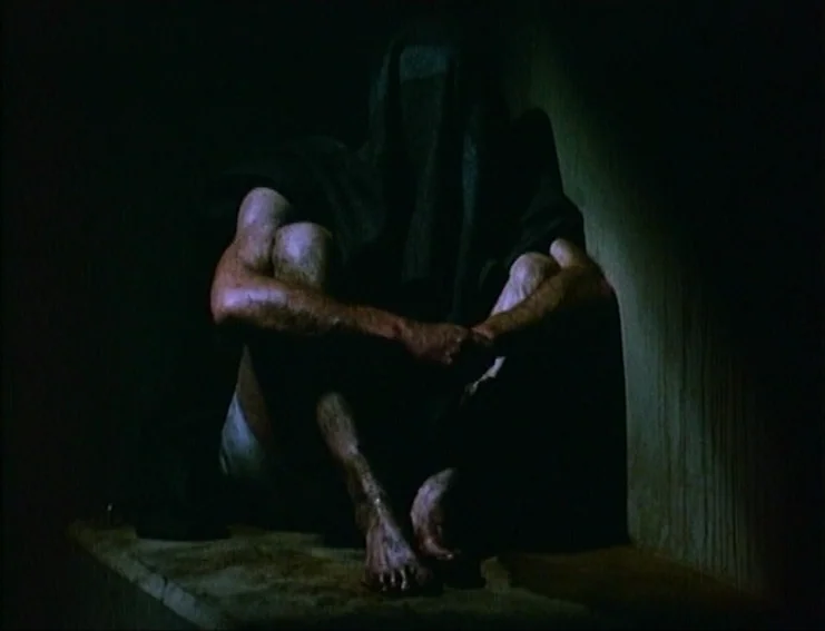 Ghosts… Of The Civil Dead (1988) movie still: medium — A dark, confined space with stone-like surfaces and no visible background detail. A cloaked…