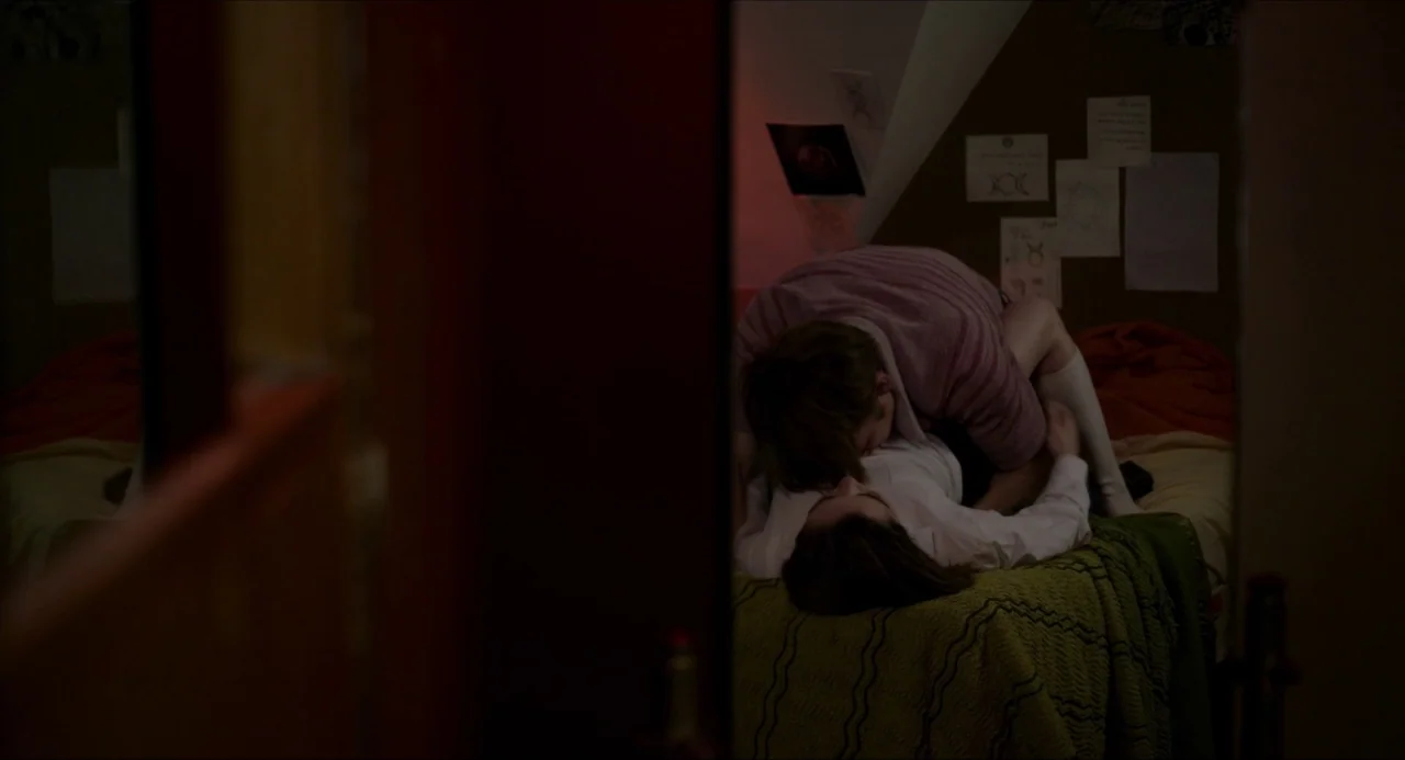 The Falling movie still: medium — A dimly lit room with walls adorned with taped drawings and notes. A man and a woman lie together on a bed covered…
