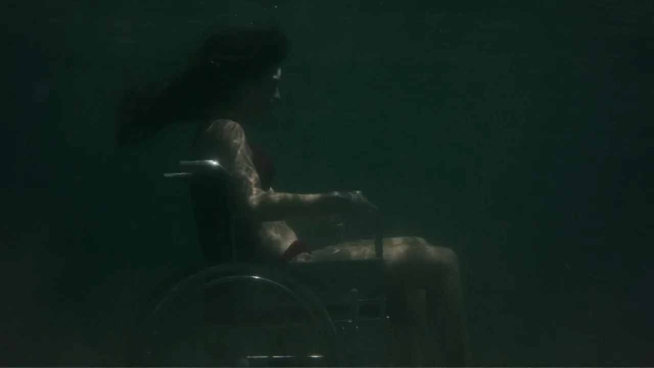 Hot Milk (2025) movie still: medium — A dark, murky body of water envelopes the frame with limited visibility. A woman with dark hair sits in a…