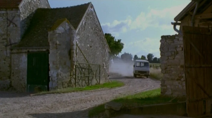 Code Unknown (2000) movie still: wide — A rural dirt road passes between two stone farm buildings in the countryside. A white van drives away along…