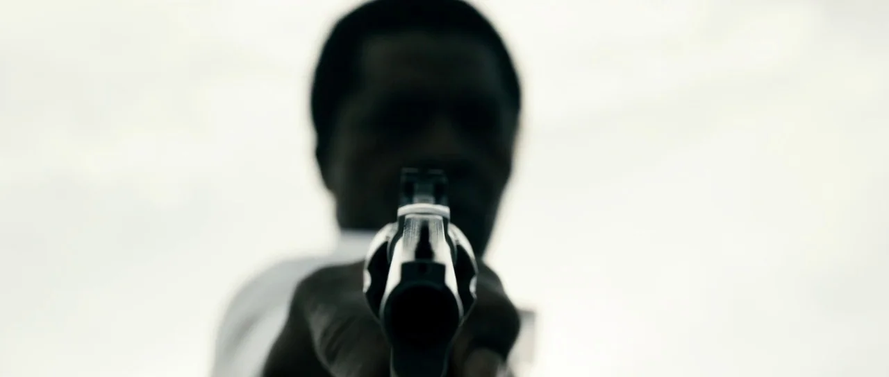 Calvary (2014) movie still: closeup — A figure stands against a bright, featureless overexposed sky. The person points a revolver directly at the…