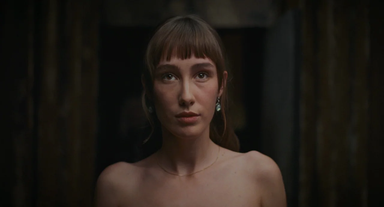 La Abuela (2021) movie still: closeup — A woman stands in a dimly lit, dark space with neutral wall textures behind her. She faces the camera…