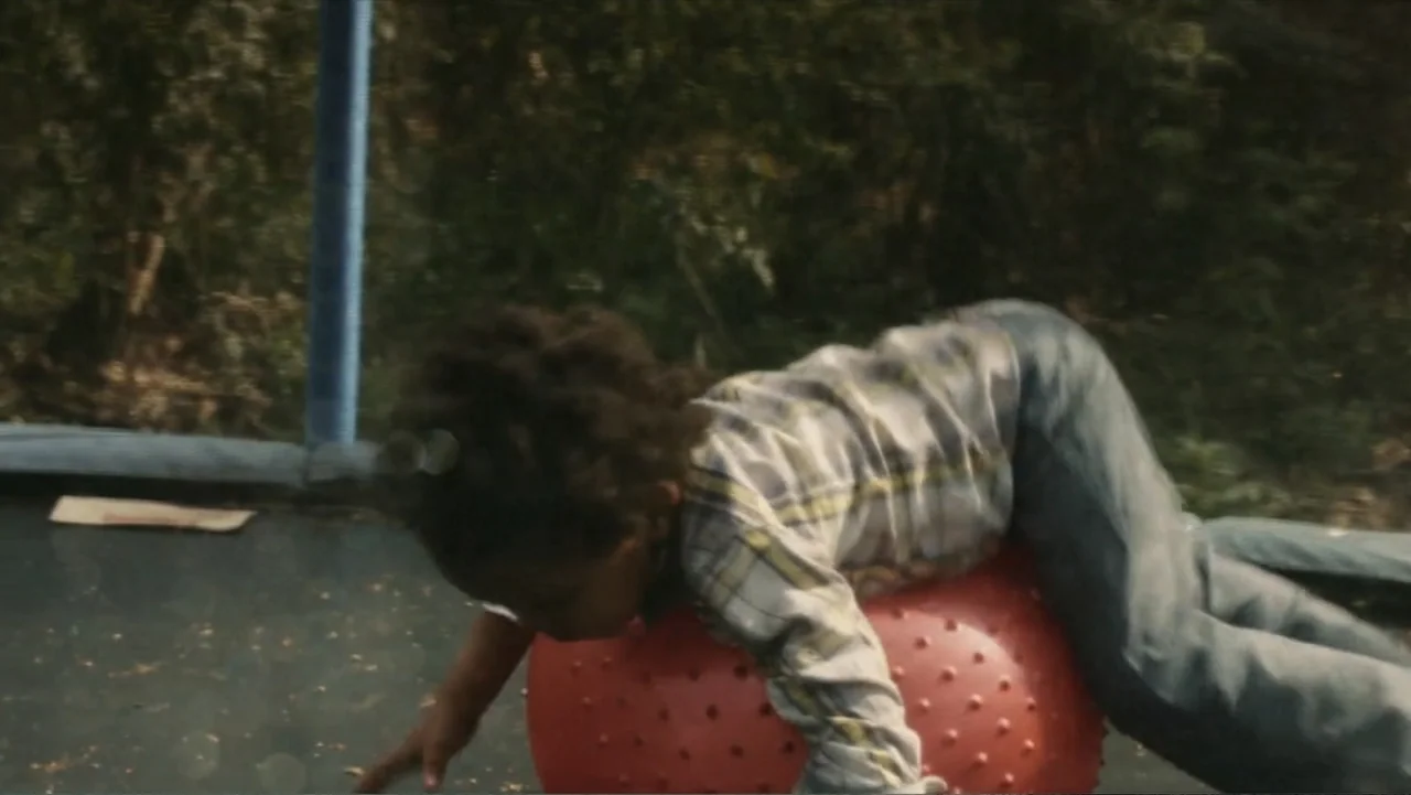 Death of a Child (2017) movie still: medium — A dark, textured trampoline surface is set outdoors amidst blurred green foliage. A child wearing a…