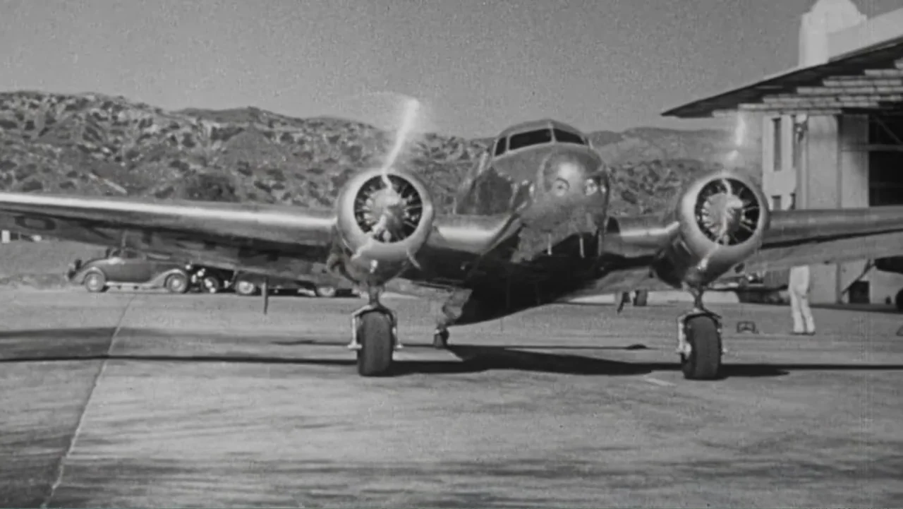 Finding Amelia (2024) movie still: wide — An airfield ramp features a silver twin-engine aircraft parked in front of a hangar. A person stands in…