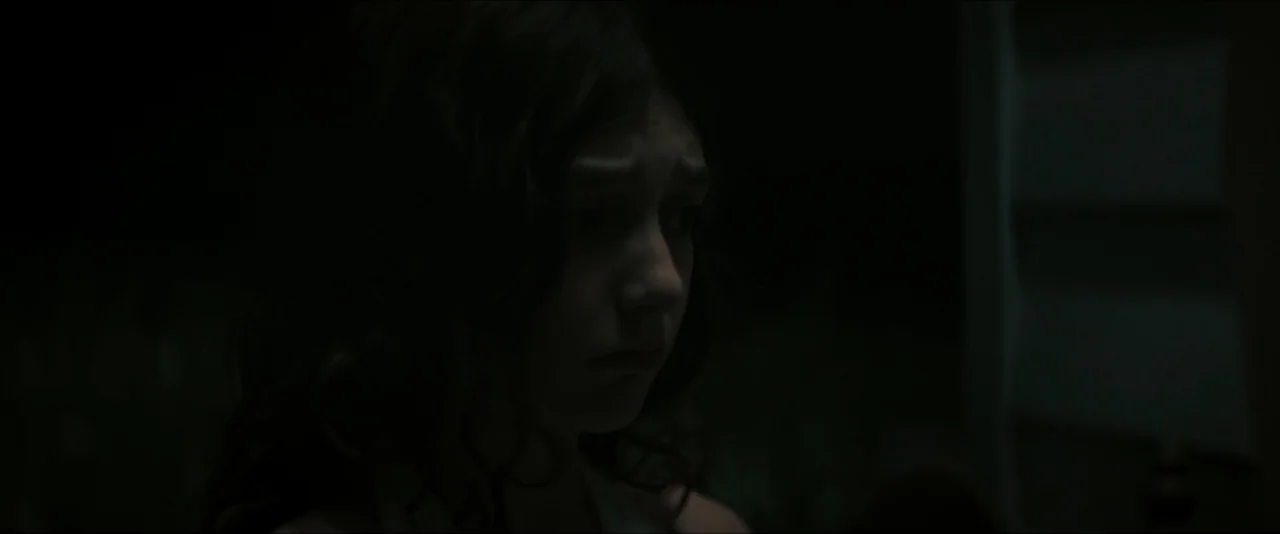 It Feeds (2025) movie still: closeup — A dark outdoor space at night near the side of a building. A girl with dark hair stands in the shadows…