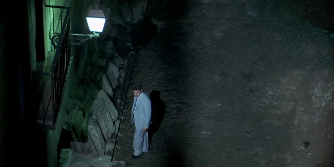 Law of Desire (1987) movie still: wide — A narrow stone-paved passage at night features a single wall with a metal balcony. A man wearing a light…