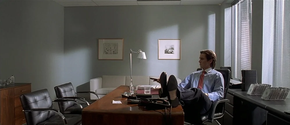 American Psycho (2000) movie still: wide — An austere corporate workspace features gray walls, minimalist furniture, and rows of compact discs. A…