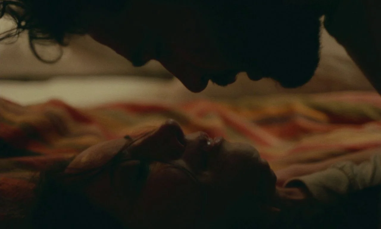 Black Bear (2020) movie still: closeup — Two figures lie facing each other on a textured, multi-colored blanket in low light. Their faces are in…