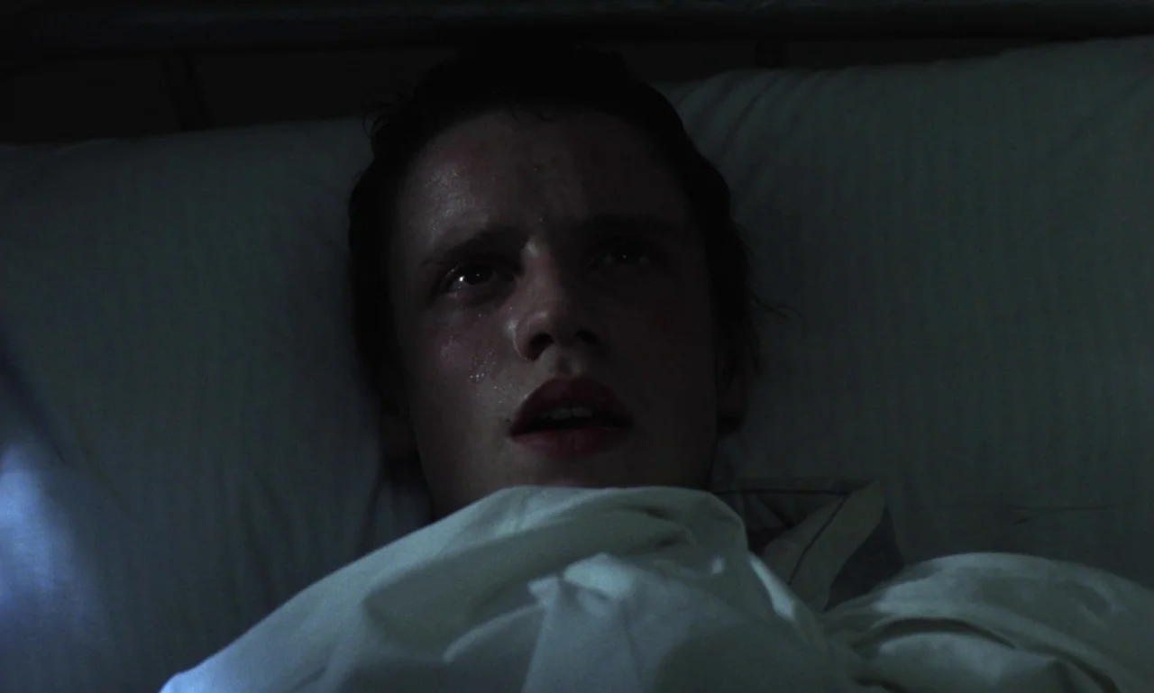 Scum (1979) movie still: closeup — A dimly lit hospital room features a person resting under white sheets. The man lies on his back with his head on…