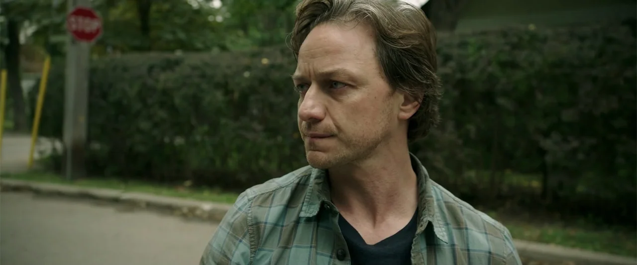K.G.F: Chapter 2 (2022) movie still: medium — A residential street with a hedge and a distant red stop sign visible. A man wearing a plaid shirt…
