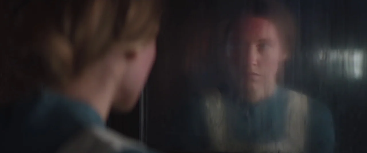 The Wind (2018) movie still: medium — A woman stares at her own reflection in a window with vertical condensation lines. The background remains…