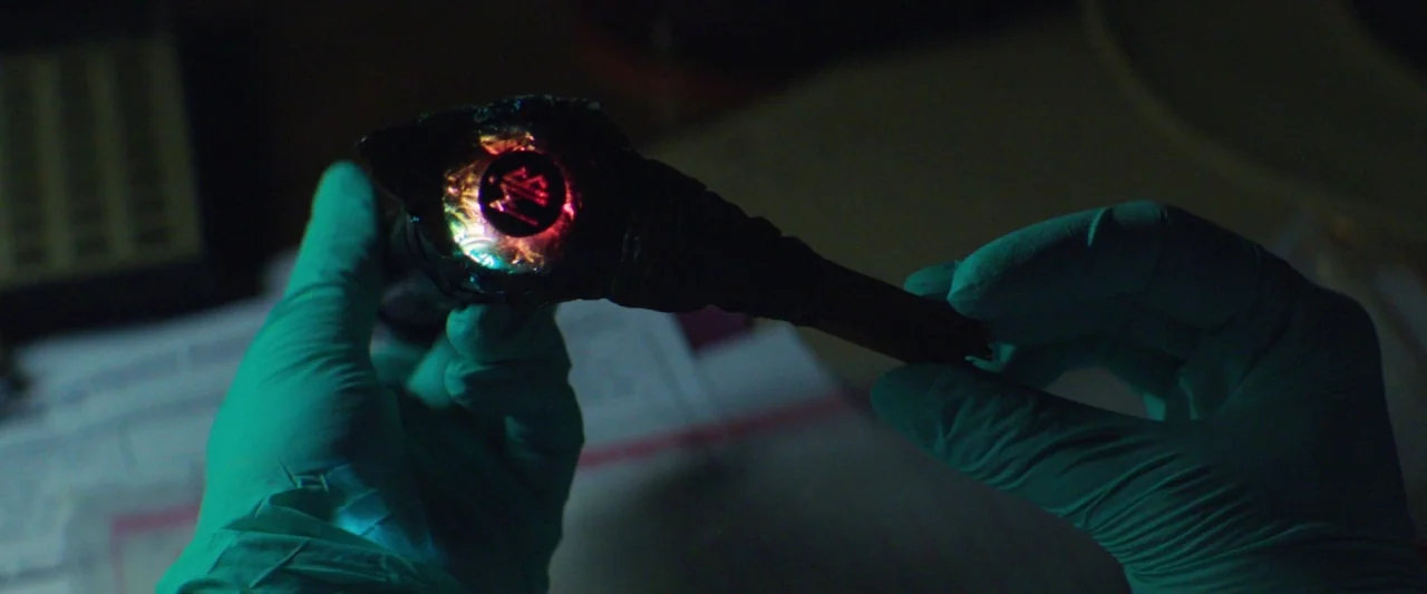 The Silencing (2020) movie still: closeup — A dark laboratory workspace contains documents under a dimly lit table surface. Two gloved hands hold a…