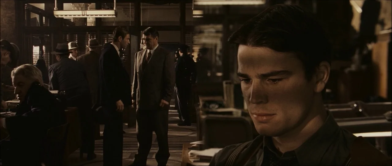 The Black Dahlia (2006) movie still: medium — A bustling 1940s-style workplace interior features multiple men in suits and fedoras at desks. A young…