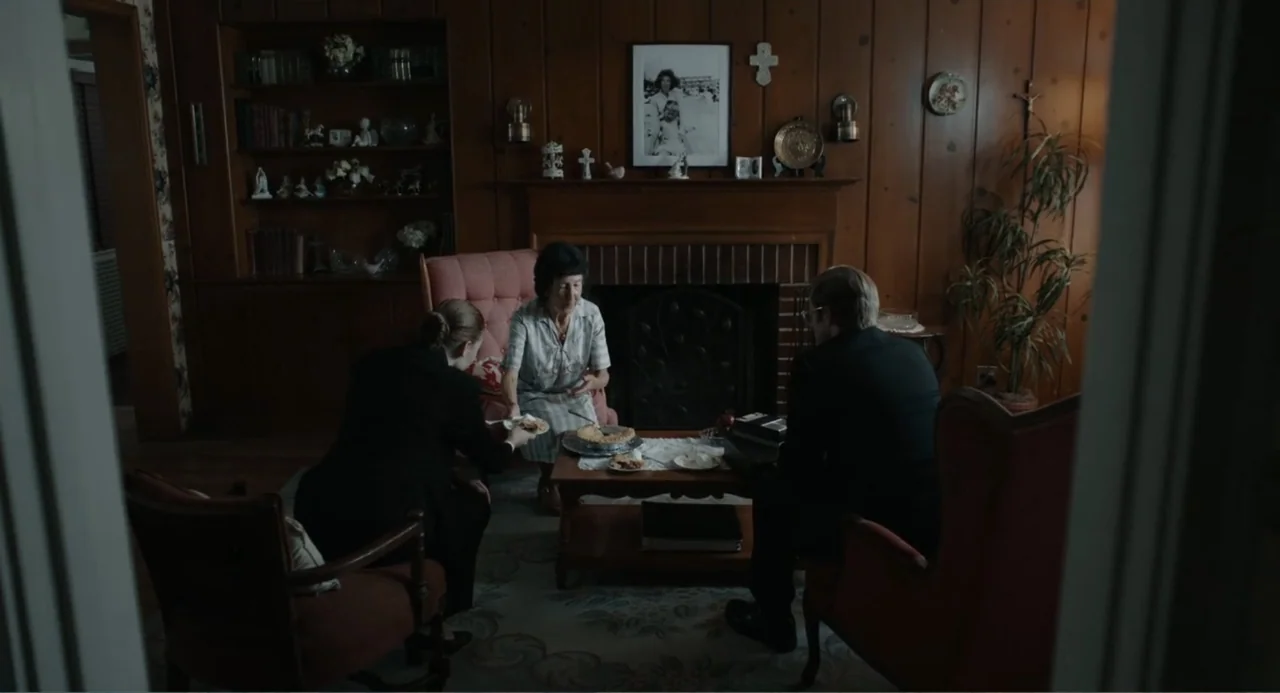 Ted Bundy: American Boogeyman (2021) movie still: medium — A 1970s living room features wood-paneled walls and a brick fireplace. Three people sit…