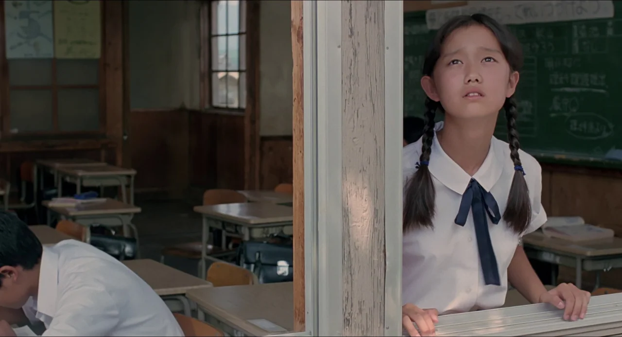 Typhoon Club (1985) movie still: medium — A school classroom features wooden desks and a large blackboard in the background. A girl sits behind a…