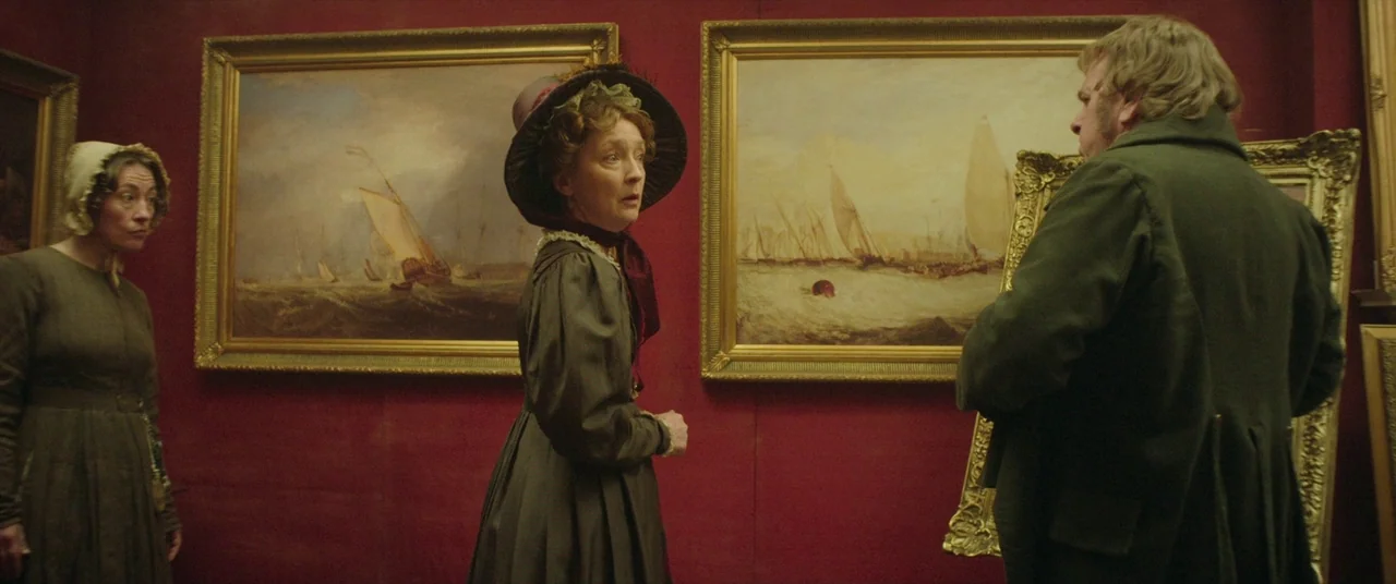 Mr. Turner (2014) movie still: medium — A gallery space with deep red walls displays several large oil paintings in ornate frames. A man holding a…