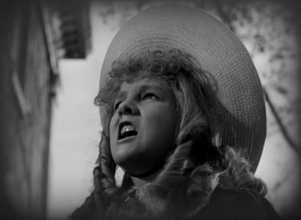 The Magnificent Ambersons (1942) movie still: closeup — A person stands outside near a building wall on a bright day. The child looks upward with an…